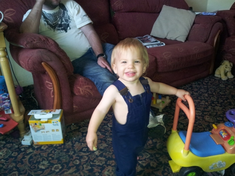 A smiling toddler with light brown hair, wearing navy blue dungarees with no shirt, stands indoors holding a yellow and orange toy walker. An adult sits on a red sofa in the background, partially out of shot, with toys and magazines nearby.