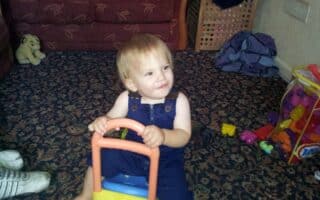 A toddler in blue dungarees sits on a yellow and orange ride-on toy, smiling and looking to the side. The child is indoors on a patterned carpet, near a maroon sofa, colourful toys, and a soft toy in the background.