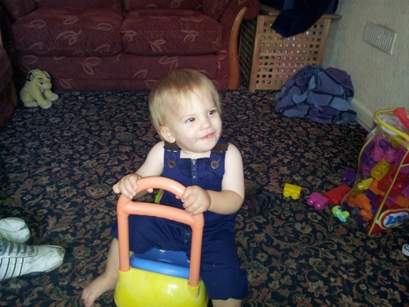 A toddler in blue dungarees sits on a yellow and orange ride-on toy, smiling and looking to the side. The child is indoors on a patterned carpet, near a maroon sofa, colourful toys, and a soft toy in the background.