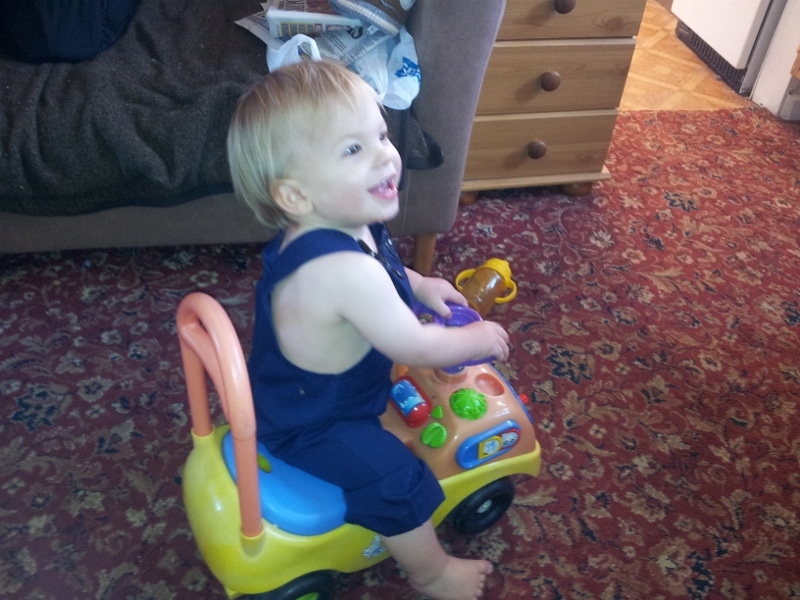A smiling toddler with blonde hair, wearing a navy blue outfit, sits on a colourful ride-on toy car indoors. The background includes a brown sofa with papers on it, a wooden chest of drawers, and a red patterned carpet covering the floor.