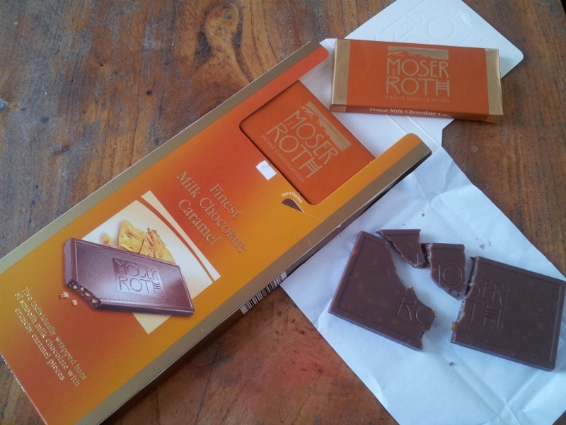 A large, opened orange box labelled “Moser Roth Finest Milk Chocolate Caramel” lies on a wooden table. Next to it are a caramel chocolate bar wrapper and a partially broken chocolate bar with “Moser Roth” embossed on the pieces.