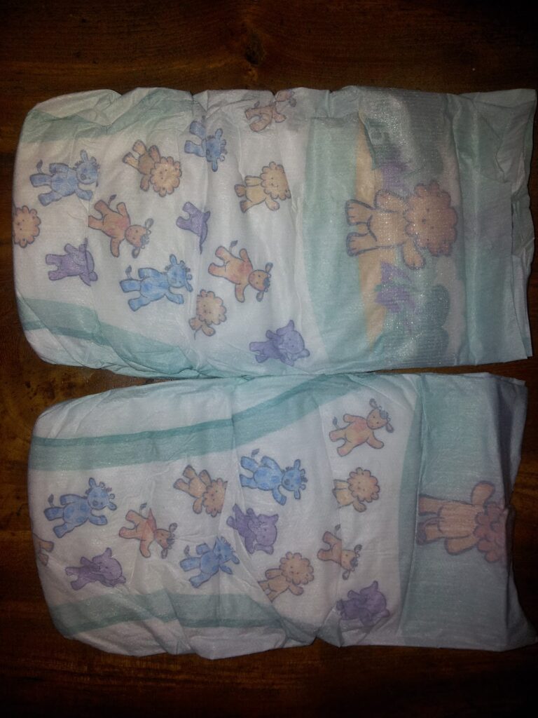 Two disposable baby nappies lie flat on a wooden surface. The nappies are white with a pastel blue-green trim and feature playful animal cartoons, such as lions, bears, and elephants, in shades of blue, purple, and orange printed across the surface.