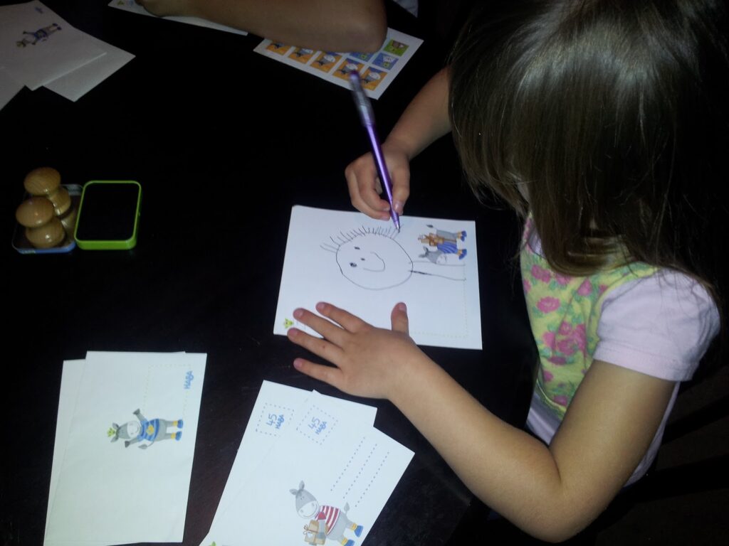 A young girl with light brown hair draws a stick figure face on white paper at a dark table. Several illustrated cards with cartoon animals, envelopes, and a green toy are scattered nearby. Only her face’s profile is partially visible.