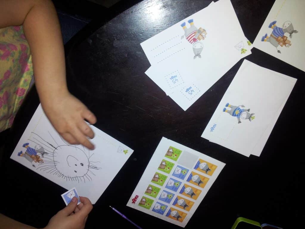 A child’s hands work on postcards at a dark table. The postcards feature cartoon animals with stamps in the corner. One postcard has a hand-drawn face and lines radiating outward. Nearby are more postcards and a sheet of colourful stamps.
