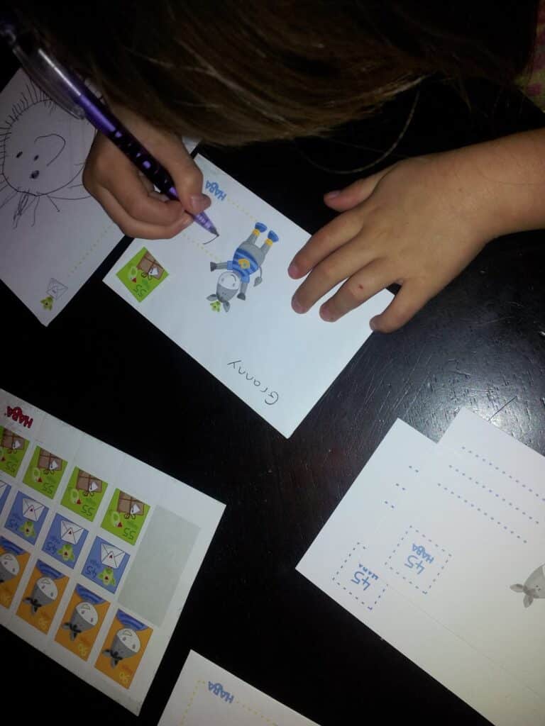 A child writes “Granny” on a card featuring a cartoon animal in uniform. Nearby are more cards and sheets with similar cartoon animal illustrations and boxes, all placed on a dark surface. The child's face is not visible.