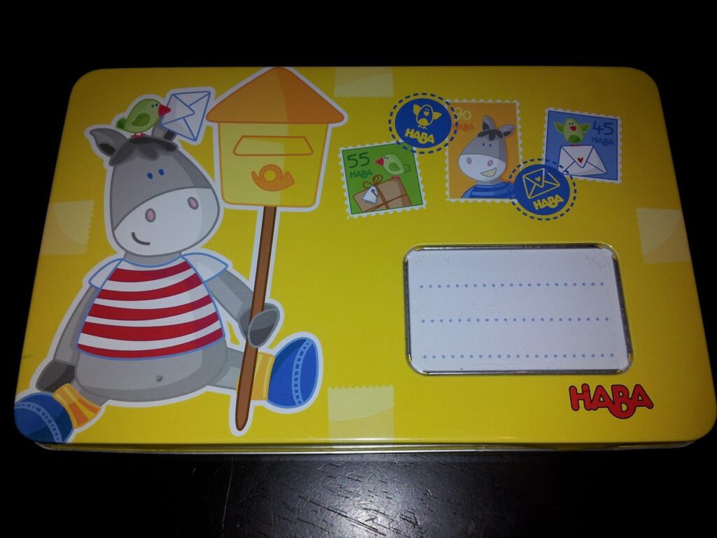 A yellow tin box features a cartoon donkey in a red-striped shirt holding a birdhouse and envelope. A green bird sits on its head. There are also three bright, playful stamp illustrations and a blank white label area, with the HABA logo in red on the bottom right-hand corner.