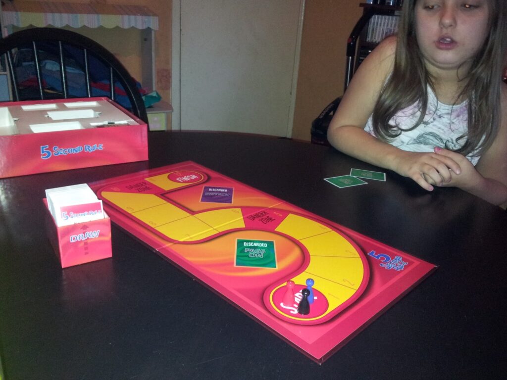 A child sits at a table playing the board game “5 Second Rule.” The game board, cards, and timer are on the table. The child holds two cards, and the game box is nearby. A bed and bookcase are visible in the background.