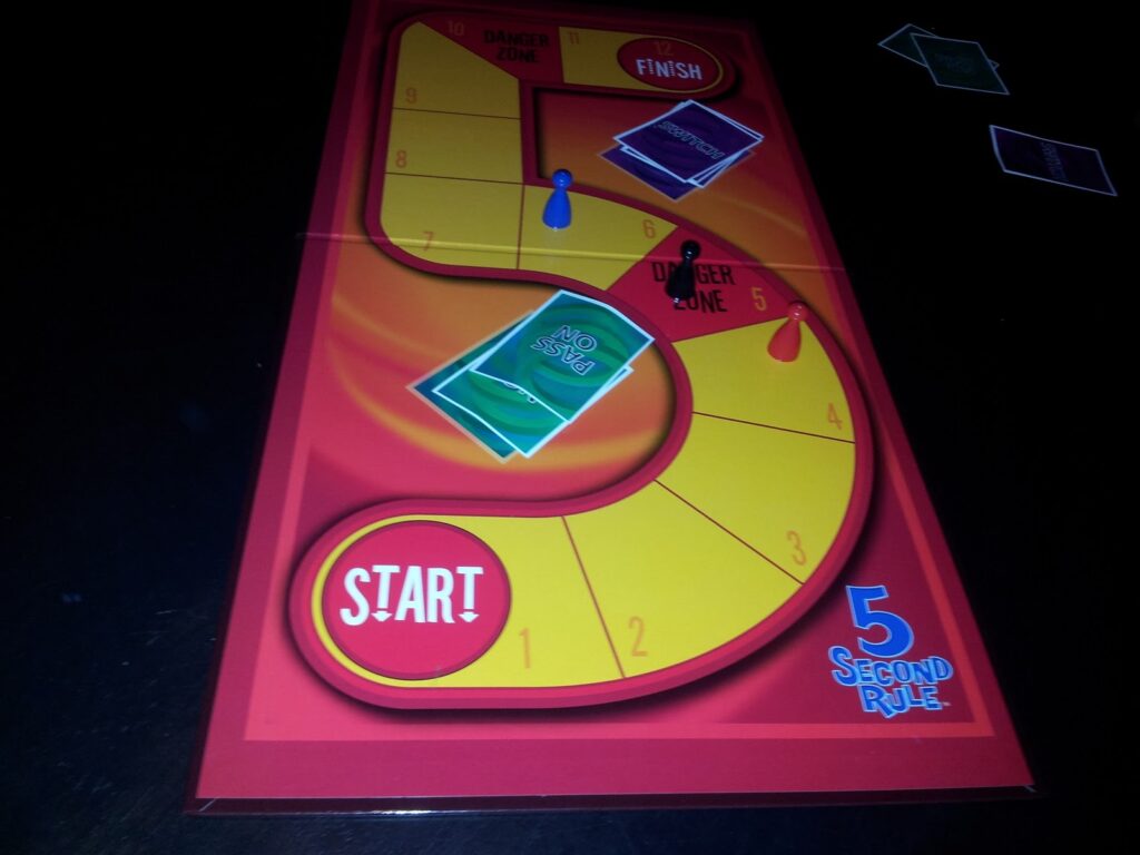 A brightly coloured “5 Second Rule” board game is shown. The board is red and yellow, shaped like an S, with spaces numbered 1–8, a “Start” circle, a “Finish” zone, and cards labelled “PASS ON” and “NO.” A blue pawn and cards are on the board.