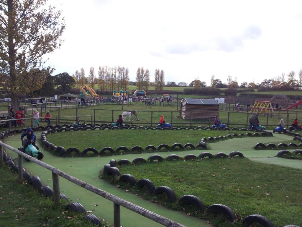 A children’s outdoor play area with kids riding small vehicles around a tyre-lined track. In the background, there are colourful playground structures, inflatable slides, and people walking or sitting. Trees and fields are visible in the distance.