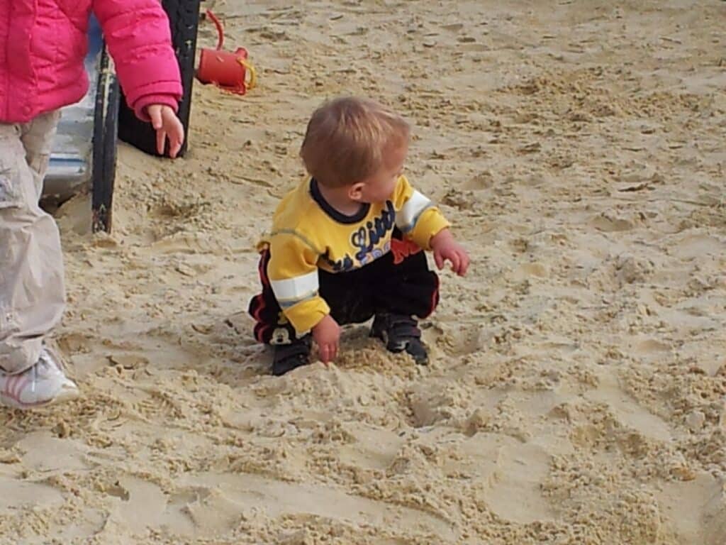 A small child in a yellow jumper and dark trousers squats on sandy ground, looking to the right. Their left hand touches the sand. An adult wearing a pink jacket and beige trousers stands nearby, visible from the hips down.