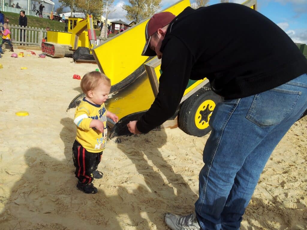 A toddler in a yellow shirt and black trousers stands on sand, smiling at an adult in jeans and a black jacket who is bending down holding something in his hands. Behind them is a bright yellow playground slide shaped like construction equipment.