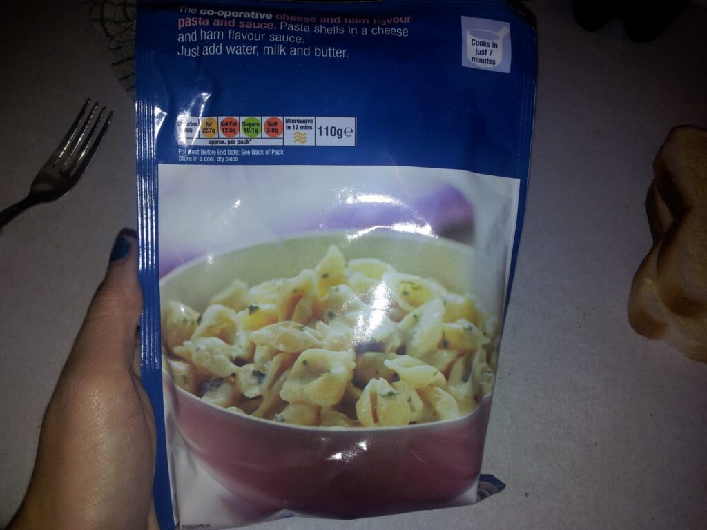 A hand holds a blue packet of The Co-operative cheese and ham flavour pasta shells. The front label shows a bowl filled with creamy pasta shells. Nearby are a fork and a slice of bread on a table. Nutrition info and cooking instructions are visible on the packet.