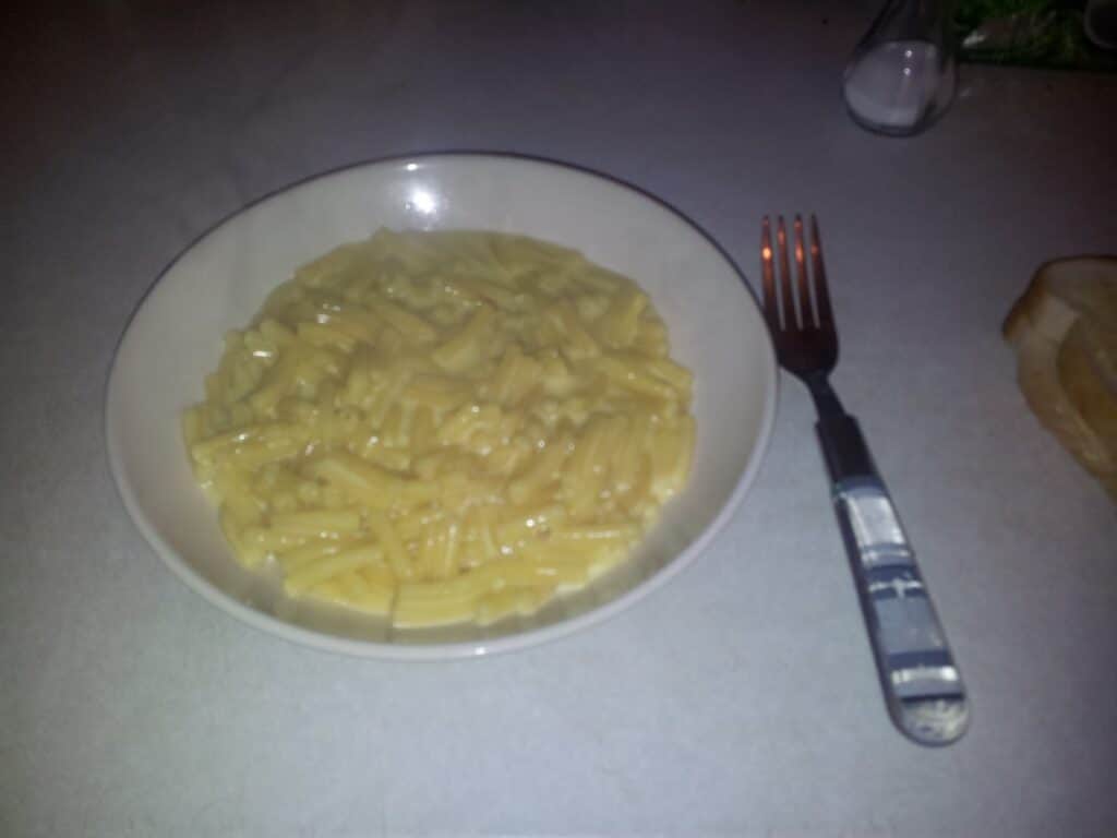 A white bowl filled with macaroni cheese sits on a light-coloured countertop. A fork with a patterned handle is placed to the right of the bowl, and part of a bread roll is visible in the top right corner. The lighting is slightly dim.