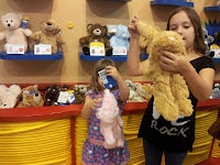 Two young girls stand in front of shelves filled with soft toys. The girl on the left holds a pink cuddly toy in front of her face, while the girl on the right, wearing a black shirt, examines a tan soft toy. Both are focused on the toys.