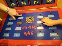 A blue board labelled “HEAR ME” features a yellow spider’s web design and multiple red buttons. An adult’s hand and a child’s hand are pressing buttons. Numbered compartments 13–16 are visible at the top of the colourful display.