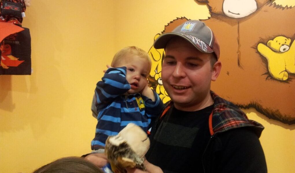 A man in a baseball cap holds a toddler in a blue striped shirt. The child covers their ears and looks unsettled. Behind them is a wall painted with large teddy bears, one brown and one yellow. A soft toy is held in the foreground.