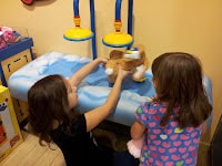 Two young girls are playing at a station with a soft toy, possibly at a Build-A-Bear Workshop. One girl holds the soft toy under blue and yellow toy “shower” fittings, while the other girl watches. The background is decorated with clouds.