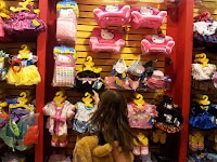 A young girl stands in front of a display wall filled with colourful doll clothes and accessories, including pink chairs, dresses, and soft toys. The wall is organised with items hanging on yellow hangers shaped like teddy bear heads.