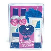 A gift set with white and blue bottles and jars labelled "Dead Sea Magik," a blue bath pouffe, and a pink card reading "I Love Dead Sea Magik Sparkling Luxury," all packaged in a clear plastic box with a pink background.