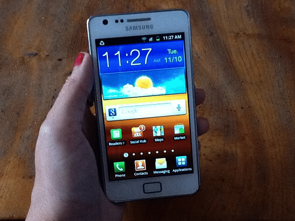 A left hand with pink-painted nails holds a white Samsung mobile displaying the home screen. The time is 11:27 am on Tuesday, 11/10. The weather widget shows a sunny sky. Several app icons are visible on the wooden table background.