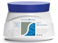 A white bath with a blue lid labelled "Dead Sea Salt Brushing." The packaging features water imagery, a grey rectangle, and text promoting minerals and exfoliation benefits. The brand appears above, and product details are printed on the side.