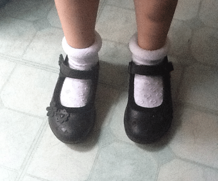 A child stands on a pale, patterned floor wearing white frilly ankle socks and black Mary Jane shoes, one of which has a flower detail. The child’s knees and lower skirt are visible at the top of the image.