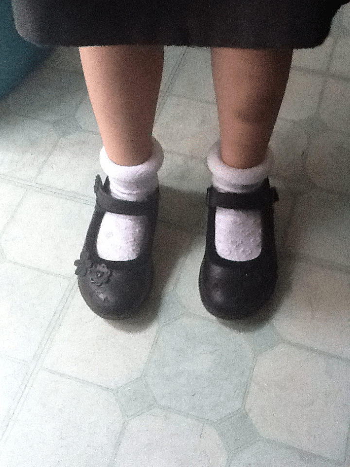 A child stands on a pale, patterned floor wearing white frilly ankle socks and black Mary Jane shoes, one of which has a flower detail. The child’s knees and lower skirt are visible at the top of the image.