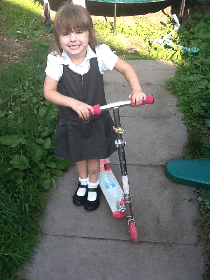 A young girl with brown hair and a fringe, wearing a white shirt, grey pinafore dress, and black shoes with white socks, stands on a garden path smiling and holding a scooter with pink handles. Green grass and a few scattered toys are in the background.