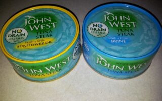 Two tins of John West Tuna Steak are shown. The left tin has a yellow rim and reads “With a little sunflower oil.” The right tin has a blue rim and reads “With a little brine.” Both feature “No Drain Less Mess” labels on white backgrounds.