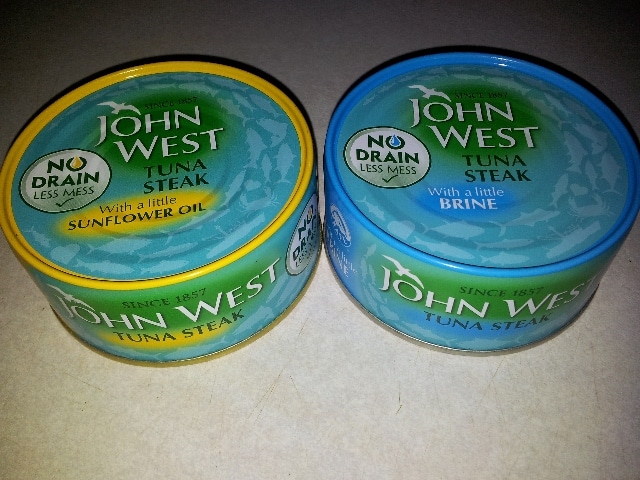 Two tins of John West Tuna Steak are shown. The left tin has a yellow rim and reads “With a little sunflower oil.” The right tin has a blue rim and reads “With a little brine.” Both feature “No Drain Less Mess” labels on white backgrounds.