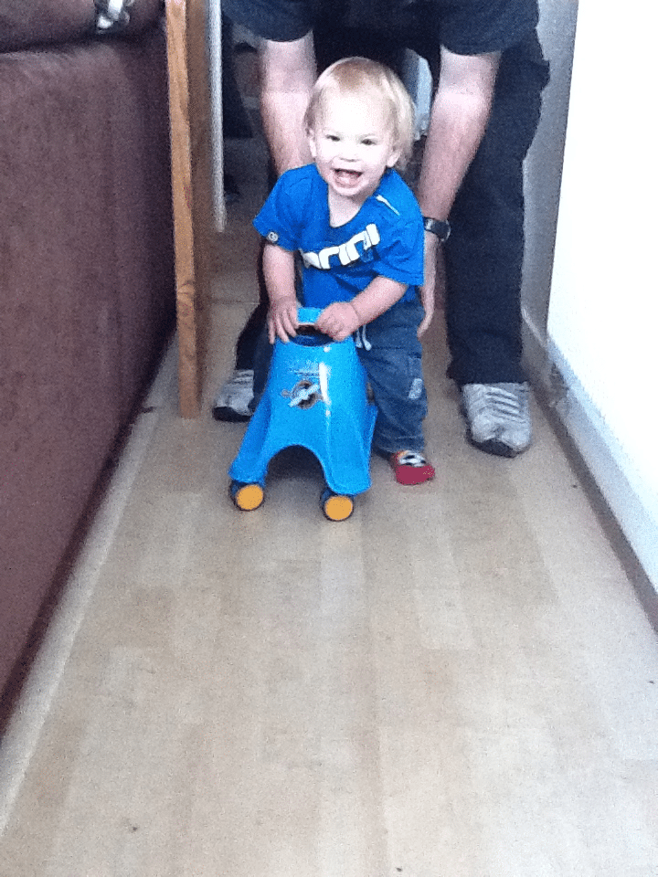 A smiling toddler in a blue shirt rides a small blue toy car indoors. An adult, visible only from the waist down, stands behind, gently holding and supporting the child. The setting appears to be a narrow hallway with light wood flooring.