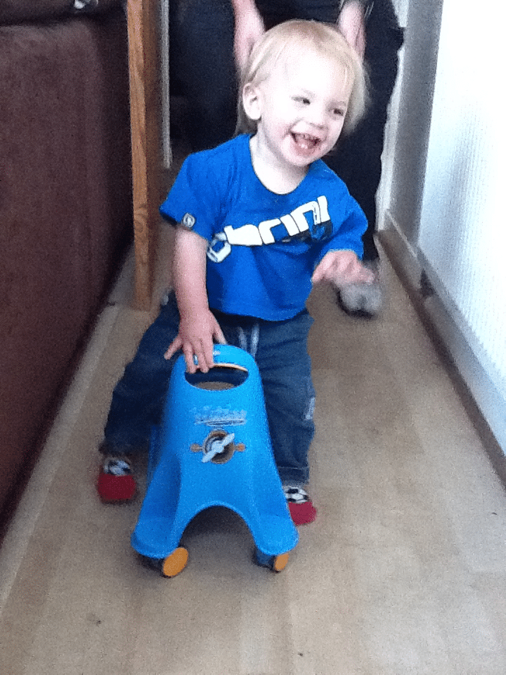 A smiling toddler with blonde hair, wearing a blue T-shirt and jeans, stands behind and holds the handle of a small blue plastic ride-on toy with yellow wheels. An adult’s hands are lightly supporting the child from behind. The setting is indoors on a wooden floor.
