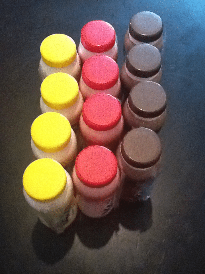 Three rows of four drink bottles seen from above, arranged in a rectangle on a dark surface. The bottles have coloured caps: yellow on the left row, red in the middle, and brown on the right, creating a visually organised pattern.