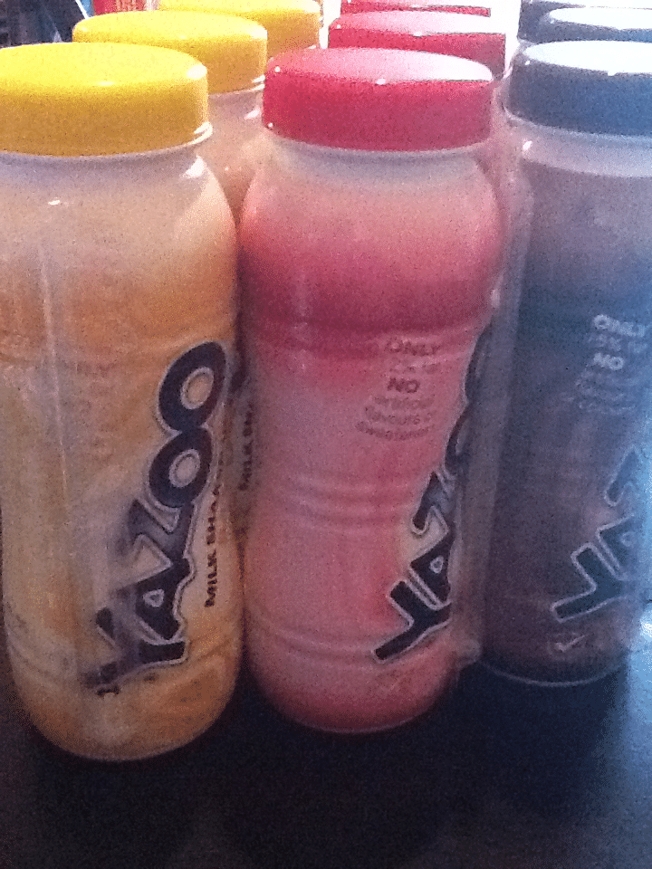 A close-up of Yazoo milkshake bottles shows three flavours: banana with a yellow cap, strawberry with a red cap, and chocolate with a brown cap. The bottles are arranged in rows on a dark surface. The brand name “Yazoo” is printed vertically.