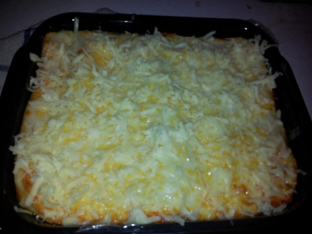 A rectangular baking tray filled with a baked casserole or pasta dish, topped with a generous layer of melted grated cheese that is golden and slightly bubbly around the edges. The dish is on a white surface.