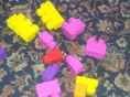 Colourful plastic building blocks, mainly yellow, pink, purple, and one red, are scattered on a patterned, floral carpet with a dark background. The blocks are different shapes and sizes, appearing randomly arranged.