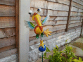 A colourful, whimsical metal bird garden ornament with googly eyes, green and blue wings, and an orange-yellow body is staked into the ground near a wooden fence and some green plants. The background includes a brick plinth and patio slabs.