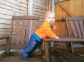 A toddler with blond hair, wearing a bright orange jacket and blue jeans, stands and leans on a wooden outdoor chair. The background features wooden cladding and a partially open wooden door. The child looks towards the camera.