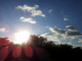 The image shows a bright sun low in a blue sky with scattered white clouds. Sunlight creates lens flares across the image. Silhouetted trees form a dark line along the horizon at the bottom of the image.