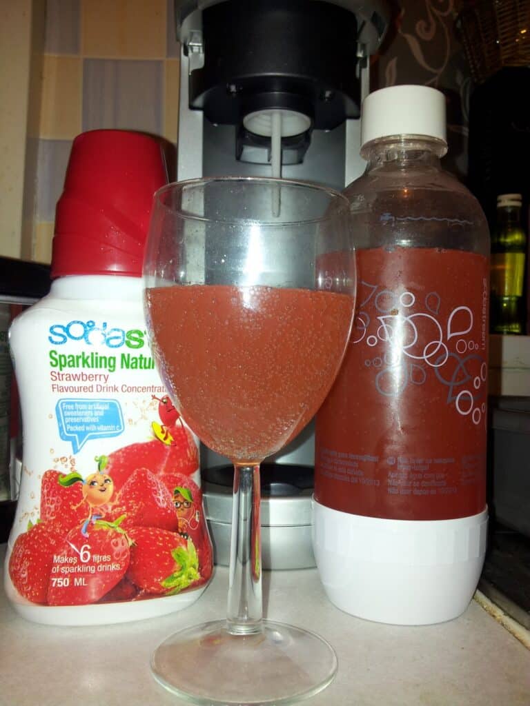 A glass of pink sparkling drink sits between a bottle of strawberry-flavoured sparkling drink concentrate and a clear plastic bottle with bubbly red liquid. A drinks machine is visible in the background. The worktop is white.