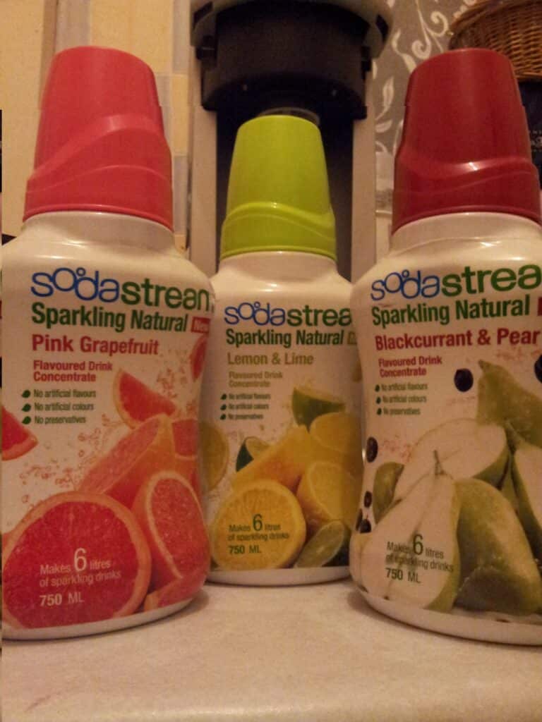 Three SodaStream sparkling drink concentrate bottles are shown: Pink Grapefruit (pink cap), Lemon & Lime (green cap), and Blackcurrant & Pear (red cap). Each bottle shows fruit images and states it makes 6 litres of drink, 750 ml per bottle.