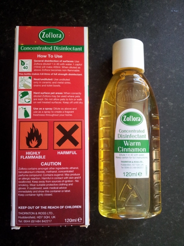 A bottle of Zoflora Concentrated Disinfectant Warm Cinnamon (120ml) sits next to its box. The box is red and green, displaying usage instructions, warnings, and hazard symbols for "Highly Flammable" and "Harmful" on the front.