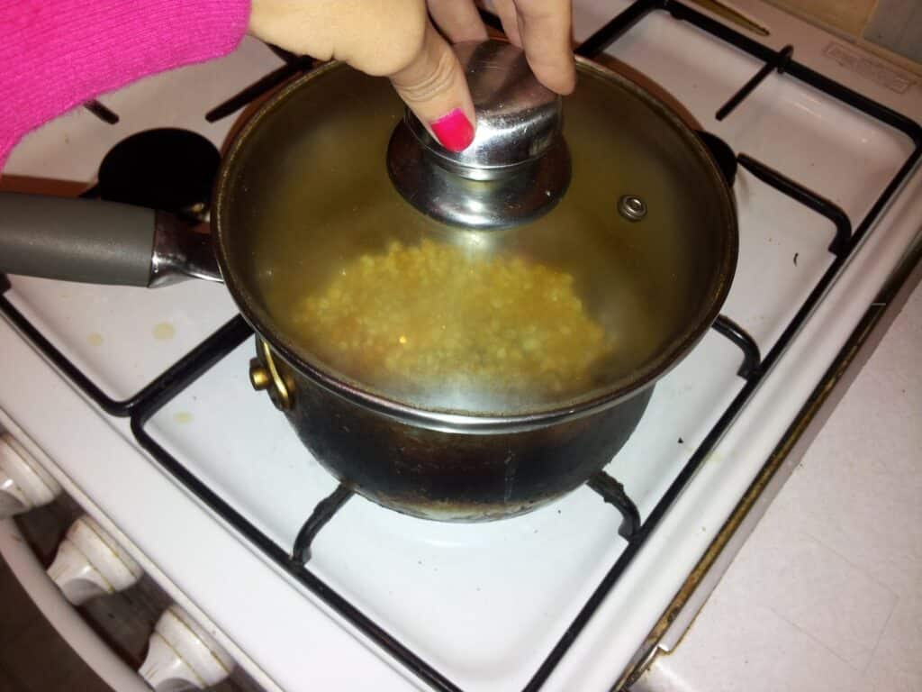 A hand with pink nail varnish holds the lid on a pot of popcorn popping on a white gas cooker. The popcorn can be seen through the glass lid as it cooks. The person is wearing a bright pink sleeve. The hob and worktop are partially visible.