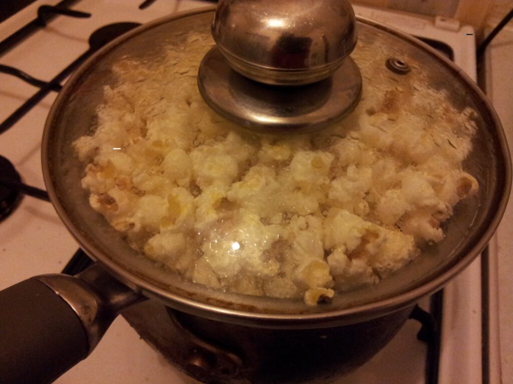 A glass-lidded saucepan on a hob is filled with freshly popped popcorn. The lid is fogged up with steam, and some popcorn is pressed against the glass. The hob burners and control knobs are visible in the background.