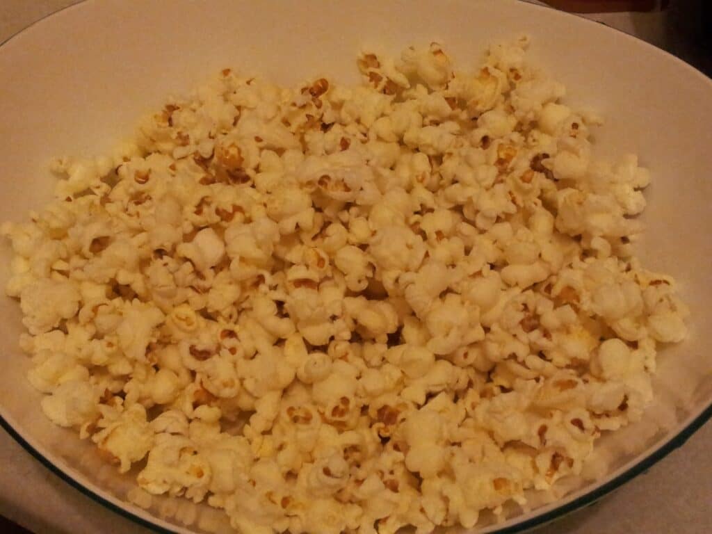 A large, white bowl filled with freshly popped popcorn sits on a light-coloured surface. The popcorn is lightly golden in some spots, with fluffy, irregular shapes. The bowl is almost overflowing, and no toppings or seasonings are visible.