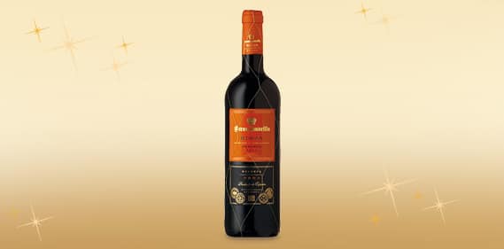 A single dark glass wine bottle with an orange foil-wrapped neck and an ornate orange label stands upright on a soft golden gradient background with scattered sparkling star effects.