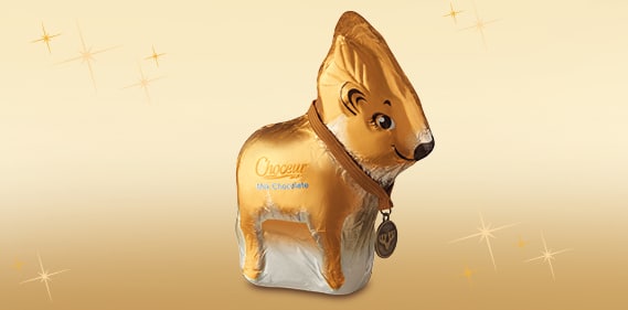 A gold-foil-wrapped chocolate bunny with a white belly stands upright. It has a brown ribbon and a round medal around its neck. The bunny is set against a light golden background with sparkles. The word “Choceur” is printed on its side in blue.