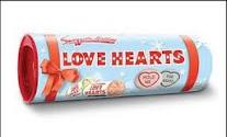 A roll of Swizzels Love Hearts sweets, wrapped in light blue packaging with a red ribbon graphic and the words "LOVE HEARTS" in bold red letters. Small colourful heart-shaped sweets are illustrated on the wrapper.