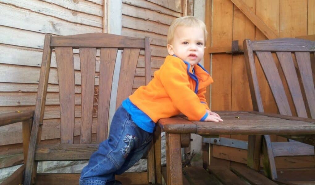 A young child in a bright orange jacket and blue jeans stands on a wooden chair, leaning forward with hands on a wooden table. The scene is outdoors, with a wooden fence and wooden gate in the background. The child looks slightly to the left.