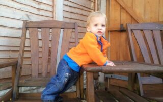 A young child in a bright orange jacket and blue jeans stands on a wooden chair, leaning forward with hands on a wooden table. The scene is outdoors, with a wooden fence and wooden gate in the background. The child looks slightly to the left.